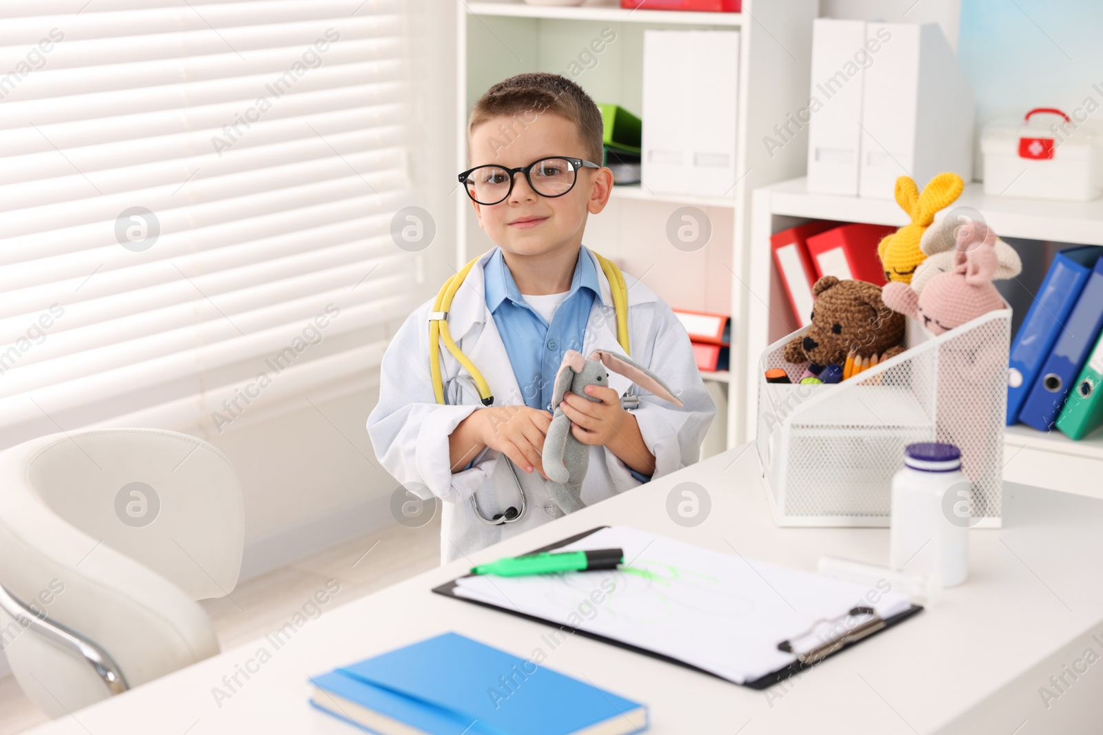 Photo of Little boy with stethoscope, toys and clipboard pretending to be doctor at white table indoors