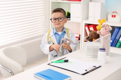 Photo of Little boy with stethoscope, toys and clipboard pretending to be doctor at white table indoors