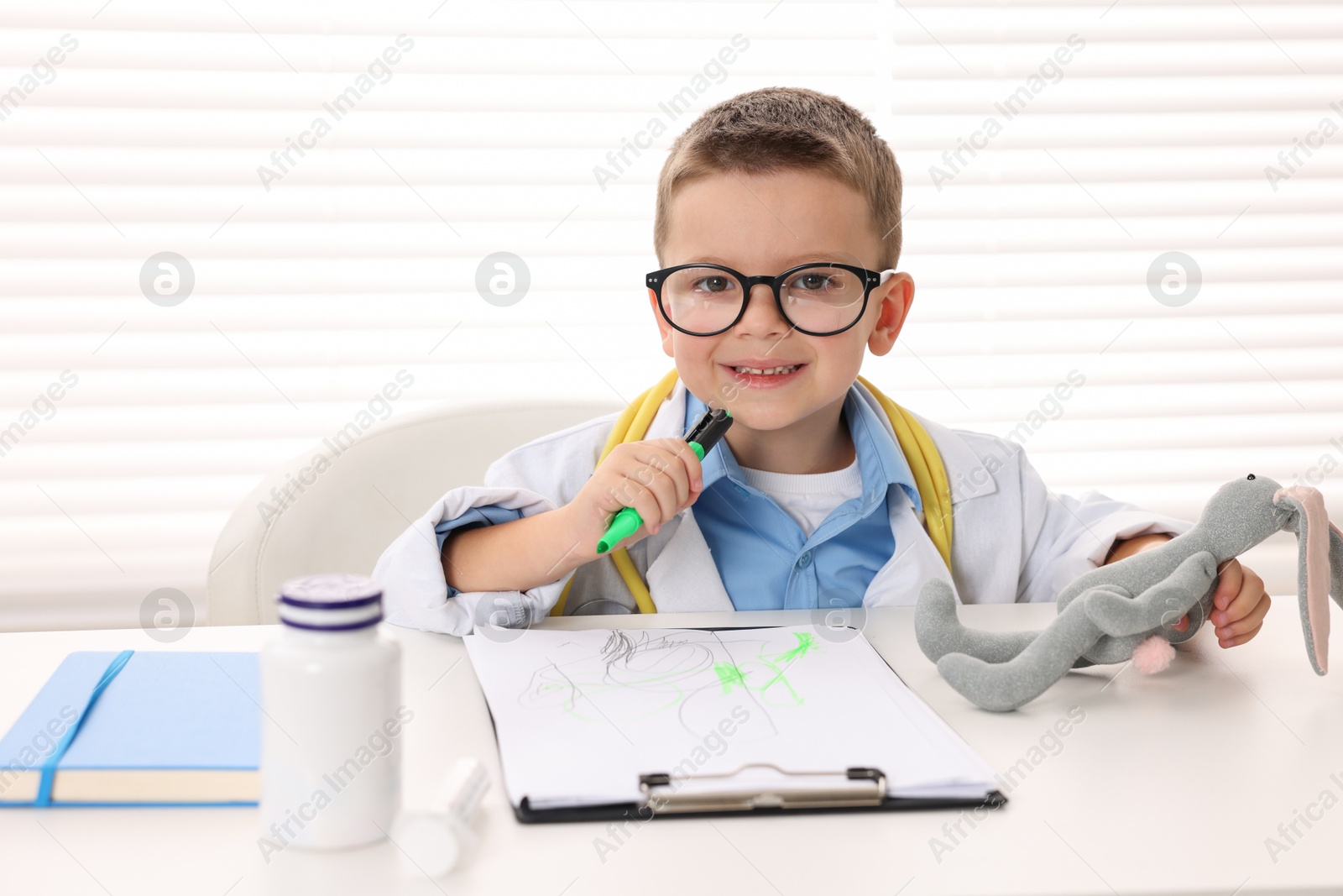 Photo of Little boy with stethoscope, toy and clipboard pretending to be doctor at white table indoors