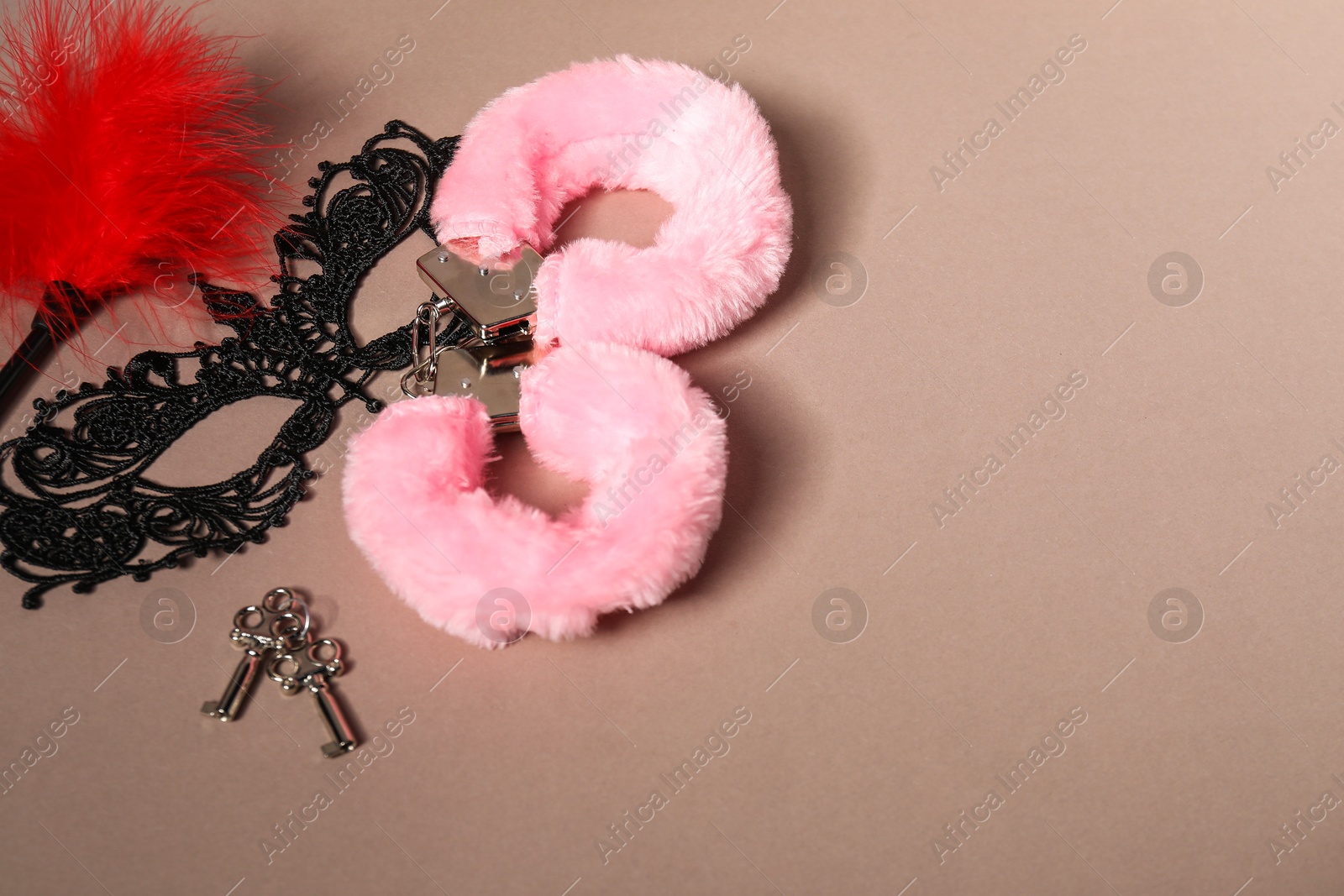 Photo of Pink fluffy handcuffs, lace mask, keys and feather on dark beige background