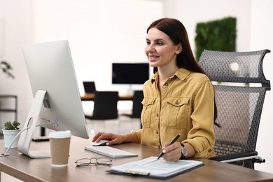 Photo of Woman taking notes while working on computer at table in office