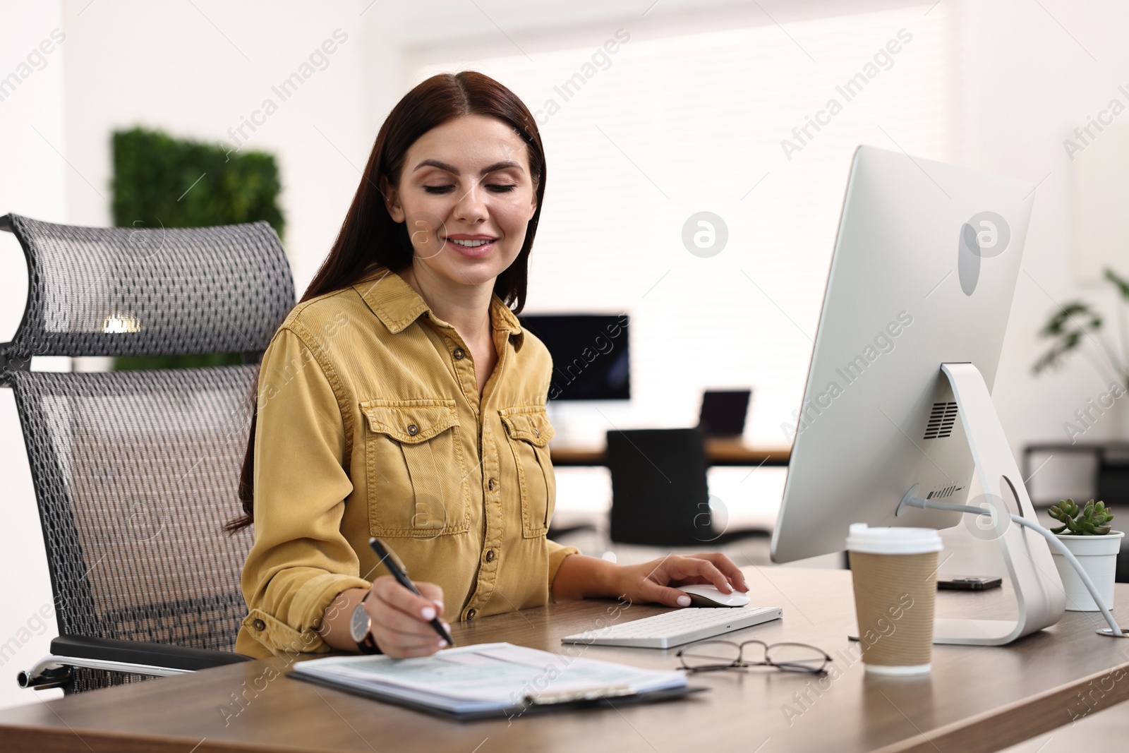 Photo of Woman taking notes while working on computer at table in office