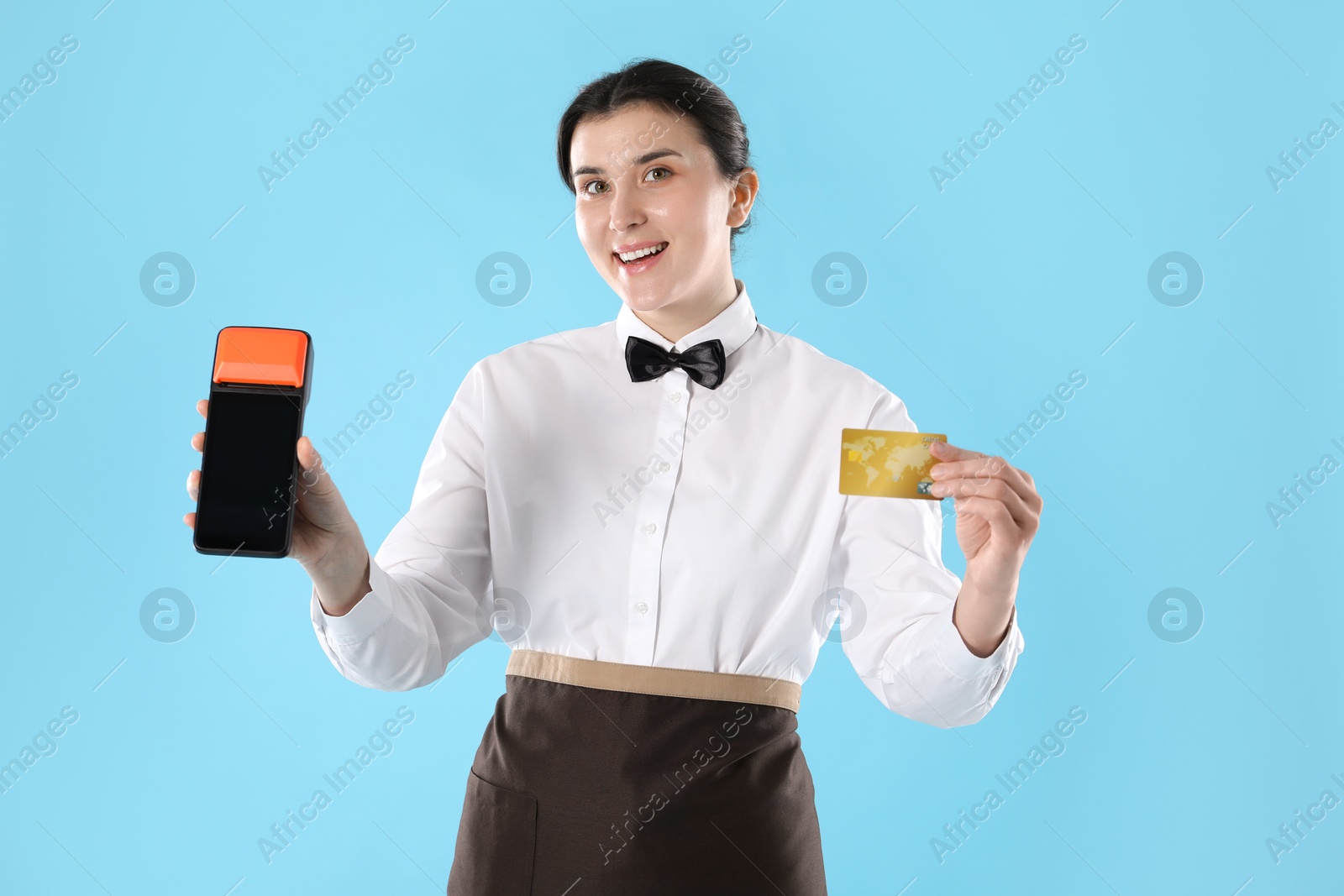 Photo of Happy waitress with payment terminal and credit card on light blue background