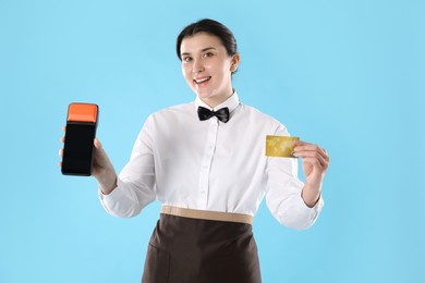 Photo of Happy waitress with payment terminal and credit card on light blue background