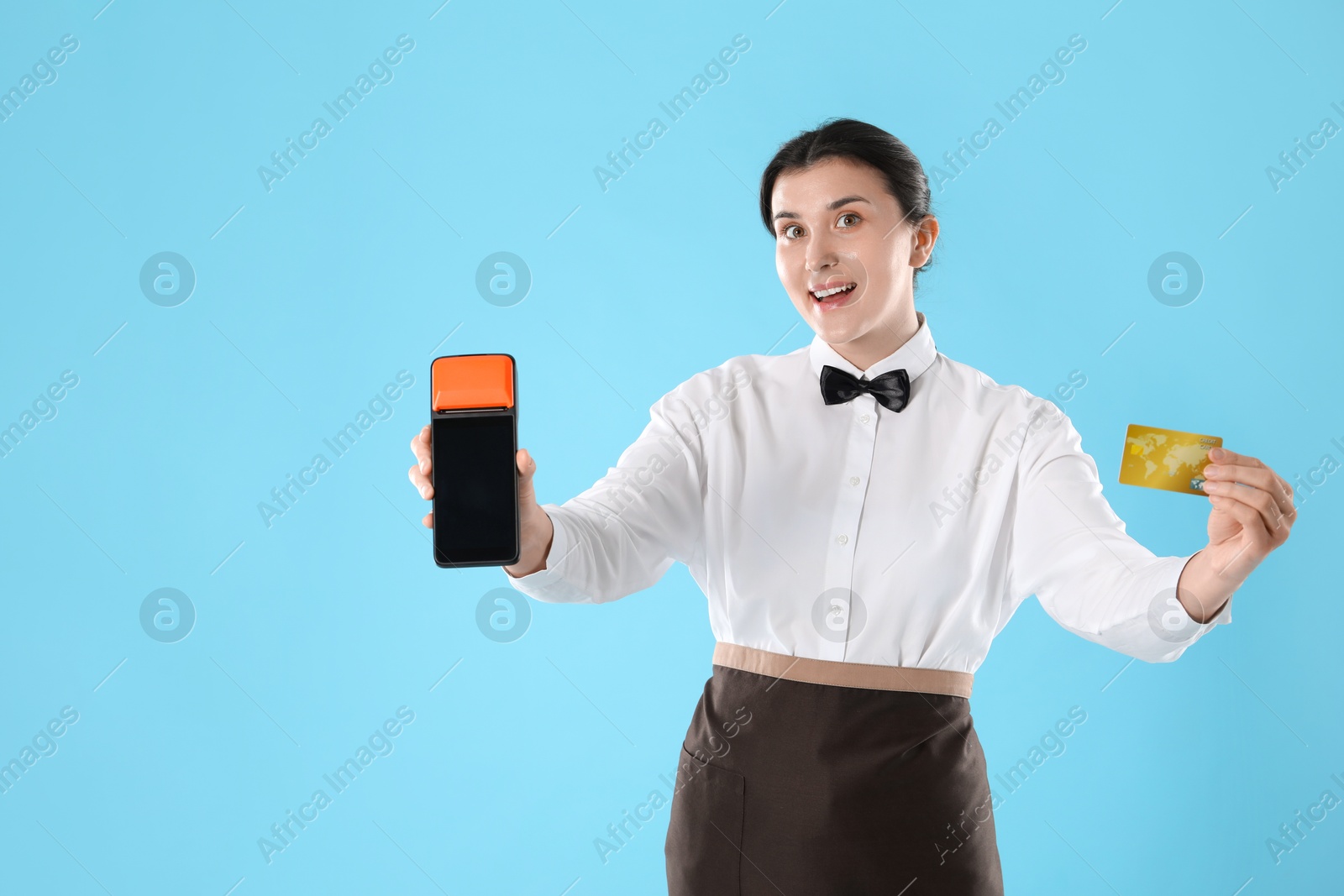 Photo of Happy waitress with payment terminal and credit card on light blue background