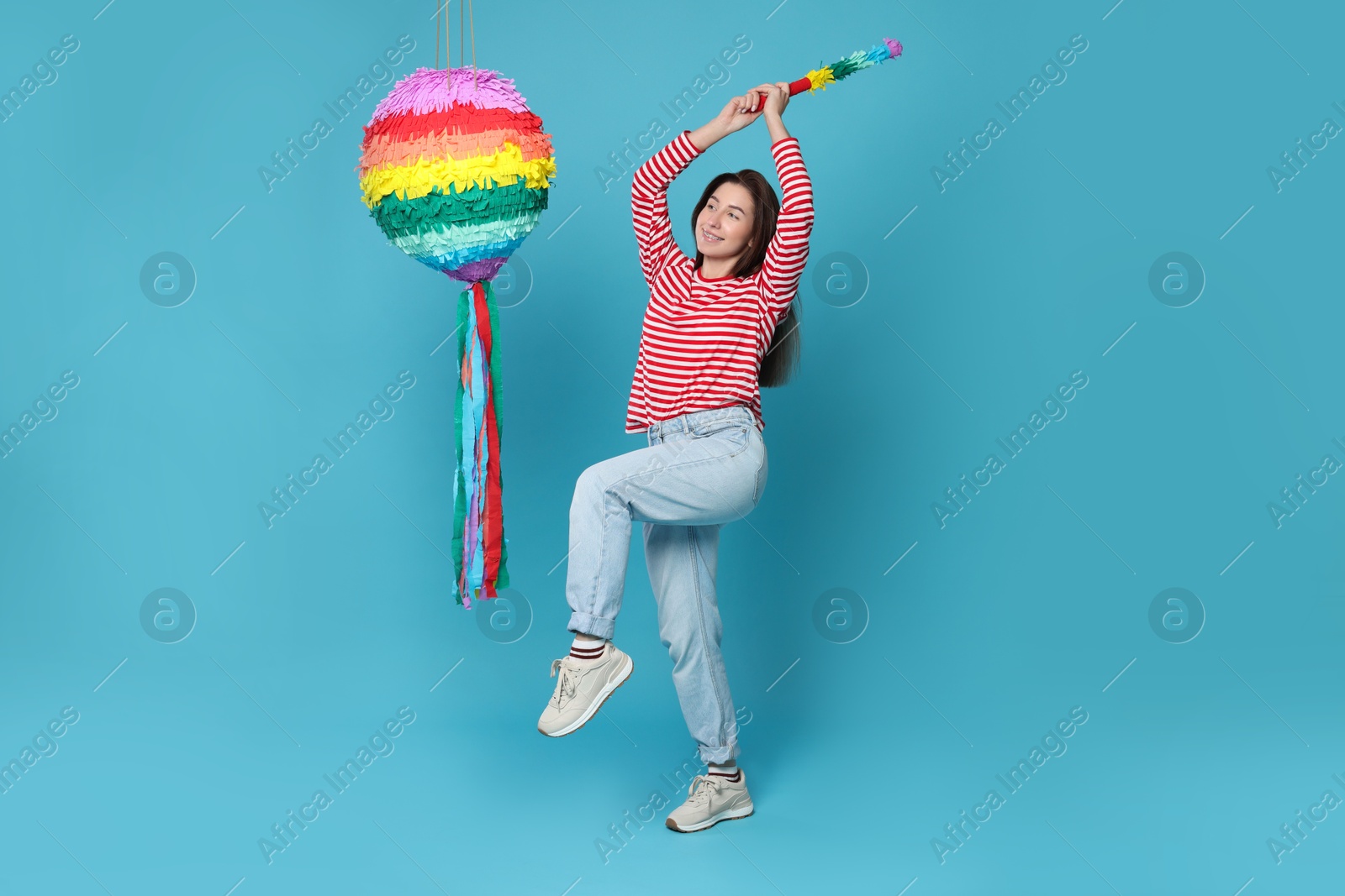 Photo of Happy woman hitting colorful pinata with stick on light blue background