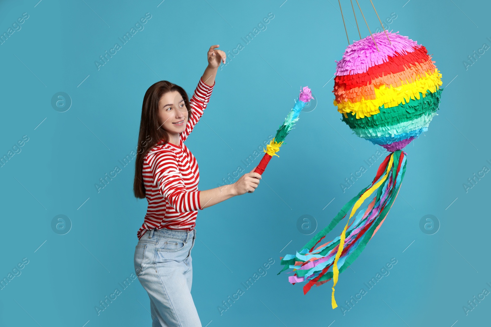 Photo of Happy woman hitting colorful pinata with stick on light blue background