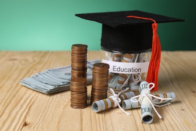 Photo of Graduate hat, dollar banknotes, coins and glass jar with word Education on wooden table, closeup. Tuition payment