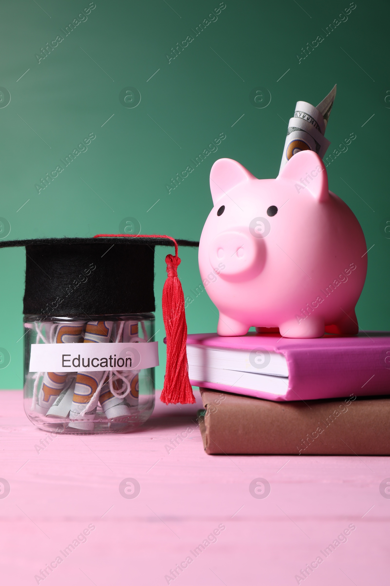 Photo of Piggy bank, graduate hat, books and dollar banknotes in glass jar with word Education on pink wooden table. Tuition payment