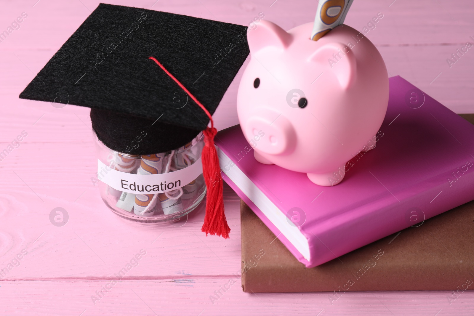 Photo of Piggy bank, graduate hat, books and dollar banknotes in glass jar with word Education on pink wooden table, closeup. Tuition payment