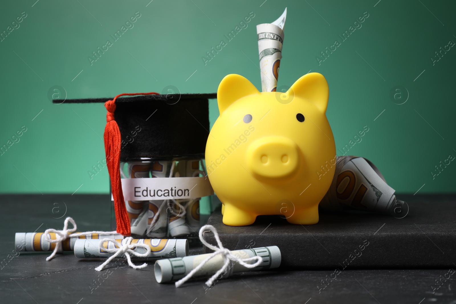 Photo of Piggy bank, graduate hat, book, dollar banknotes and glass jar with word Education on black table, closeup. Tuition payment