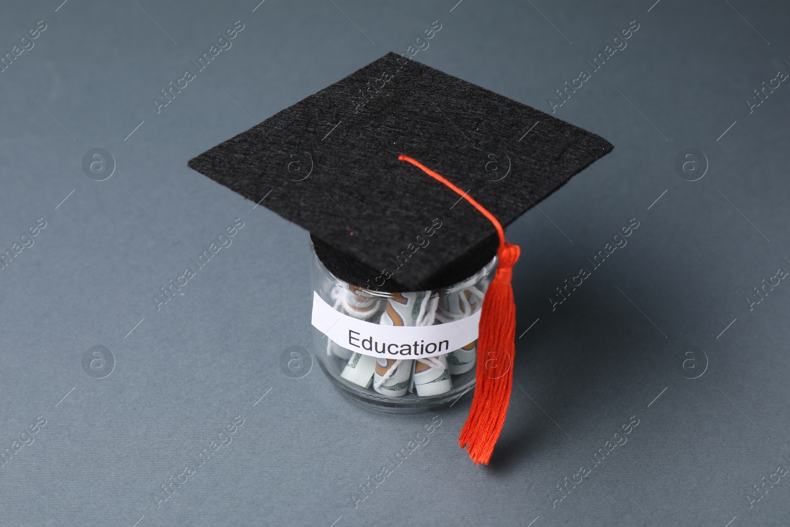 Photo of Graduate hat and dollar banknotes in glass jar with word Education on grey table, closeup. Tuition payment