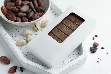 Photo of Tasty Dubai chocolate bar in package with pistachios, knafeh and cocoa beans on white textured table, closeup. Space for text