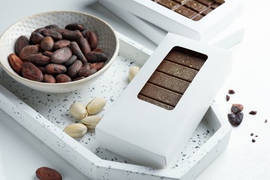 Tasty Dubai chocolate bar in package with pistachios, knafeh and cocoa beans on white textured table, closeup. Space for text