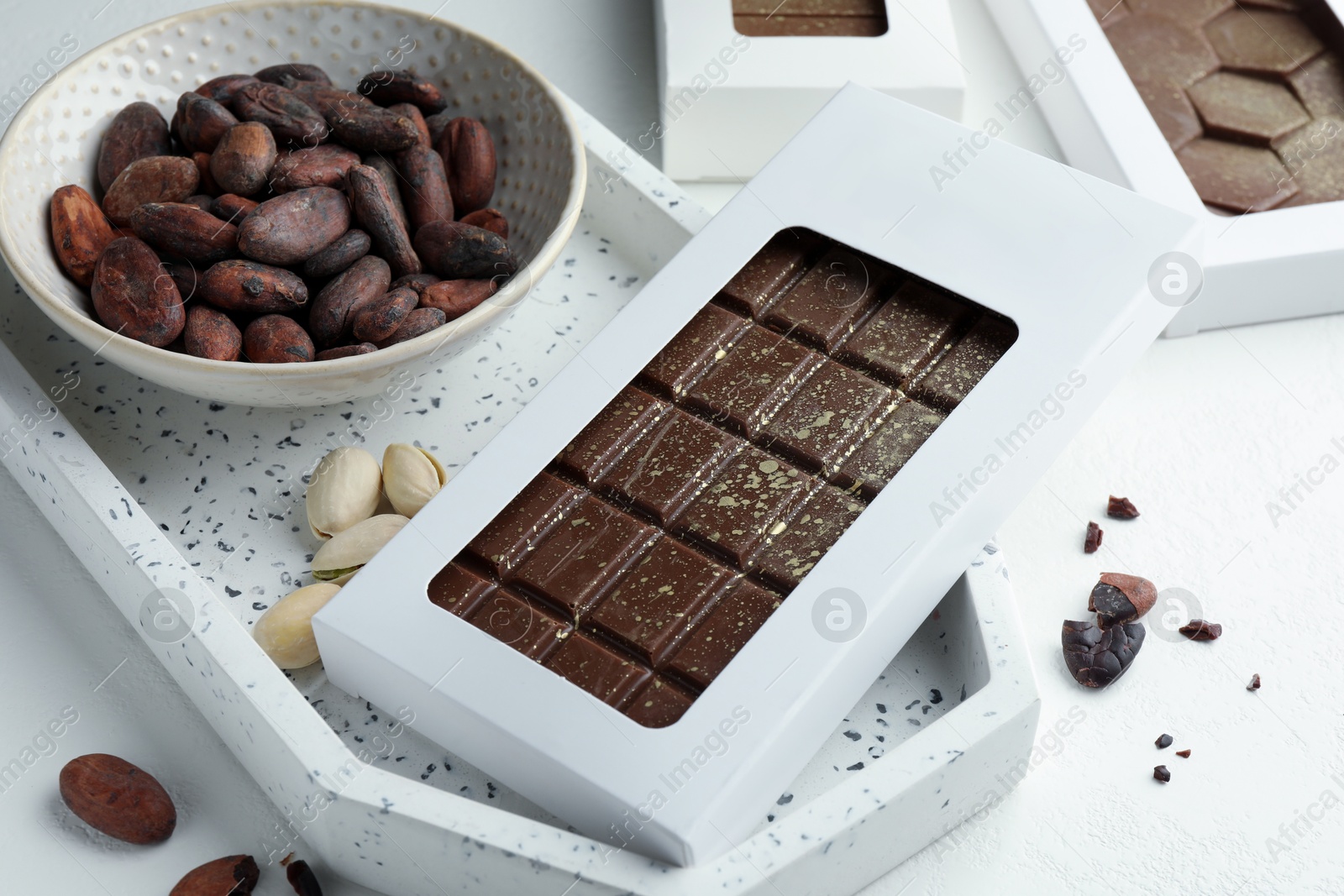 Photo of Tasty Dubai chocolate bar in package with pistachios, knafeh and cocoa beans on white textured table, closeup. Space for text