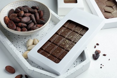 Photo of Tasty Dubai chocolate bar in package with pistachios, knafeh and cocoa beans on white textured table, closeup. Space for text