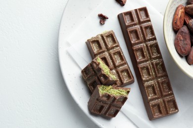 Tasty Dubai chocolate bars with pistachios, knafeh and cocoa beans on white textured table, top view. Space for text