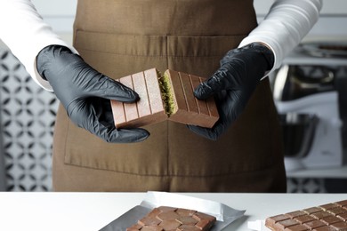 Photo of Woman breaking tasty Dubai chocolate with pistachios and knafeh at light table indoors, closeup