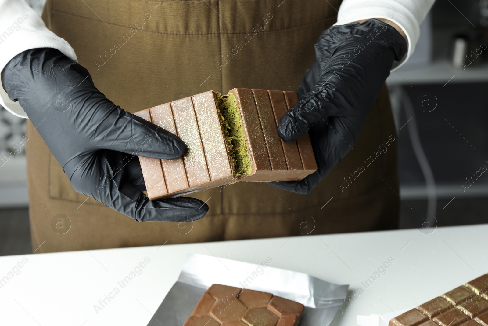 Photo of Woman breaking tasty Dubai chocolate with pistachios and knafeh at light table indoors, closeup