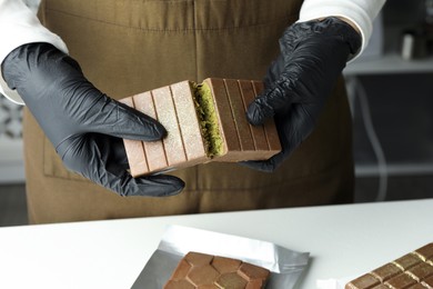 Woman breaking tasty Dubai chocolate with pistachios and knafeh at light table indoors, closeup