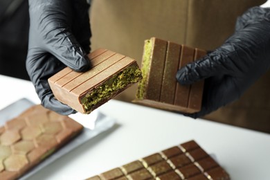 Photo of Woman holding pieces of tasty Dubai chocolate with pistachios and knafeh at white table, closeup