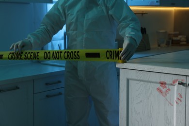 Photo of Forensic expert blocking way to crime scene with yellow tape indoors, closeup