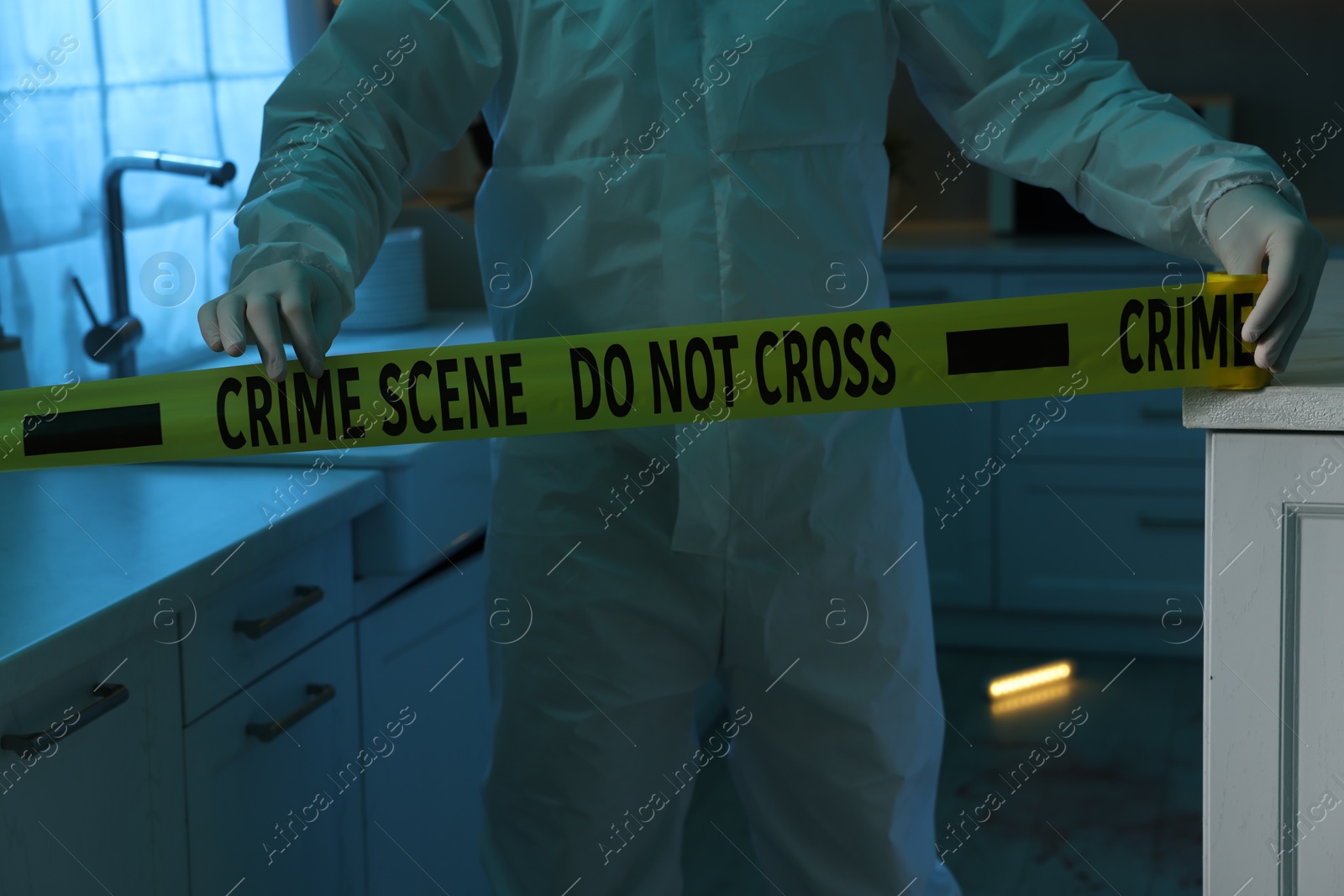 Photo of Forensic expert blocking way to crime scene with yellow tape indoors, closeup