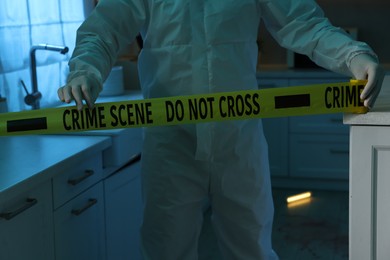 Photo of Forensic expert blocking way to crime scene with yellow tape indoors, closeup