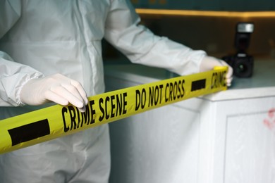 Photo of Forensic expert blocking way to crime scene with yellow tape indoors, closeup