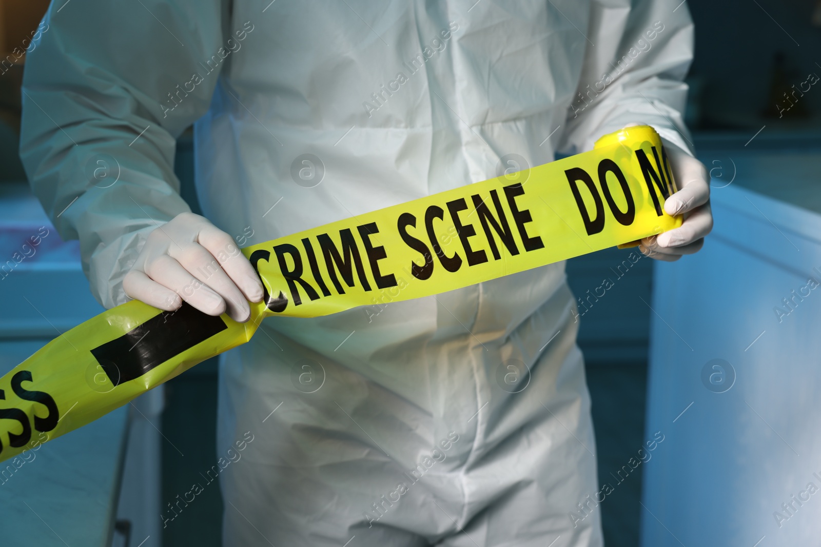 Photo of Forensic expert blocking way to crime scene with yellow tape indoors, closeup