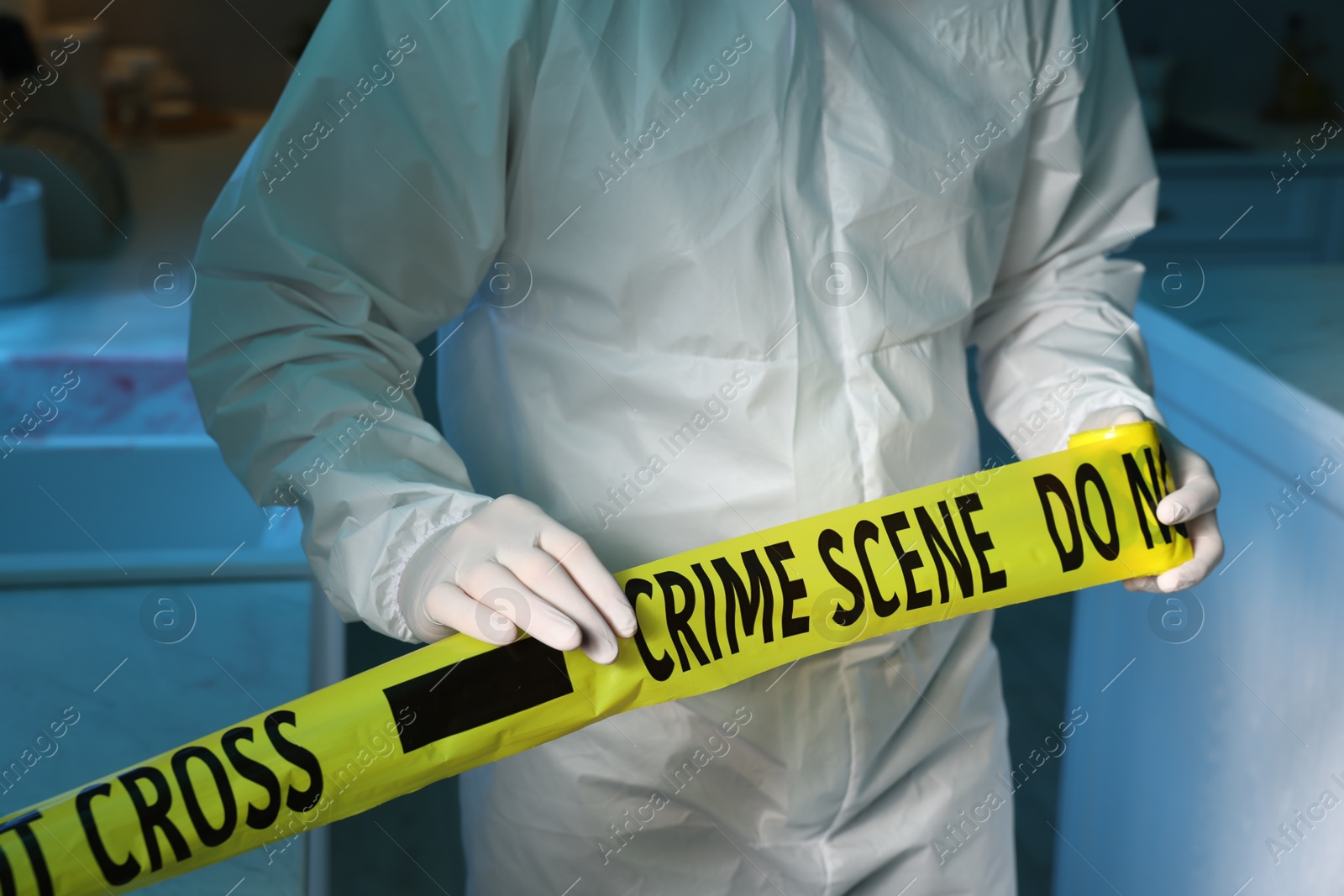 Photo of Forensic expert blocking way to crime scene with yellow tape indoors, closeup