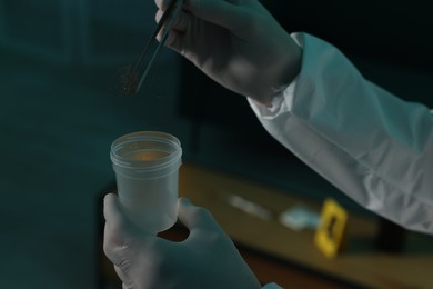 Photo of Forensic expert with tweezers collecting hair into container indoors, closeup