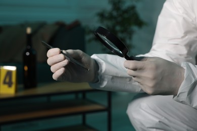 Photo of Forensic expert with tweezers examining hair through magnifying glass indoors, closeup