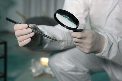 Forensic expert with tweezers examining hair through magnifying glass indoors, closeup