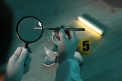 Photo of Forensic expert with tweezers examining hair through magnifying glass at crime scene indoors, closeup