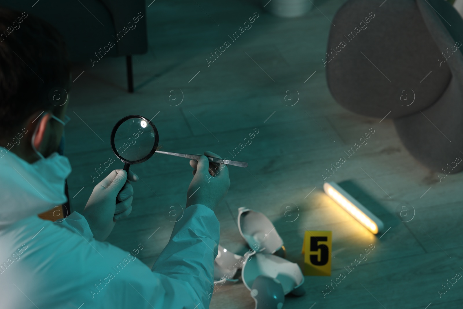 Photo of Forensic expert with tweezers examining hair through magnifying glass at crime scene indoors