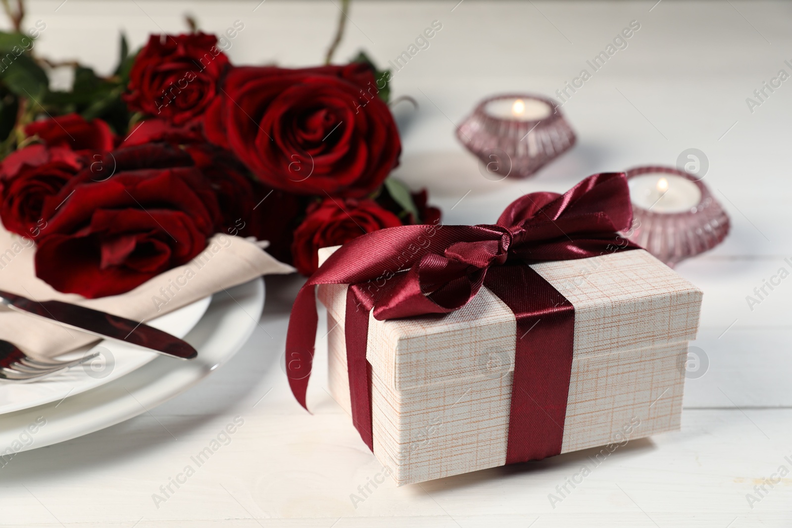 Photo of Beautiful table setting with romantic gift, flowers and burning candles on white wooden background, closeup