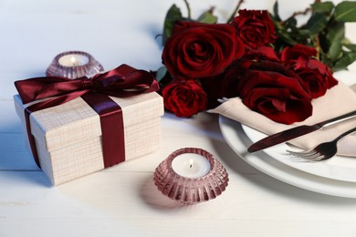 Beautiful table setting with romantic gift, flowers and burning candles on white wooden background, closeup