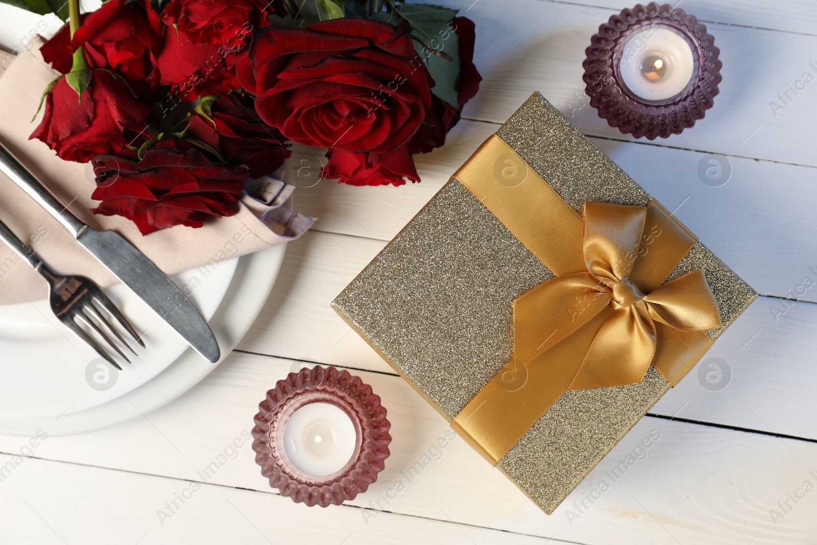 Photo of Beautiful table setting with romantic gift, flowers and burning candles on white wooden background, flat lay