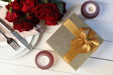 Beautiful table setting with romantic gift, flowers and burning candles on white wooden background, flat lay