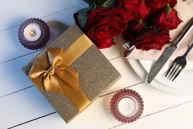 Beautiful table setting with romantic gift, flowers and burning candles on white wooden background, flat lay