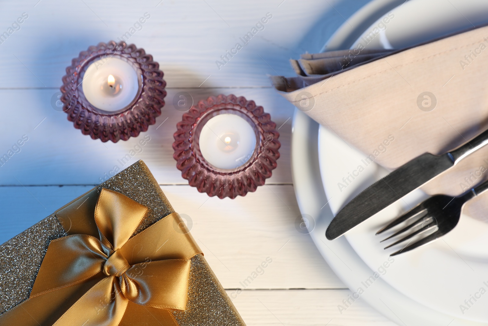 Photo of Beautiful table setting with romantic gift and burning candles on white wooden background, flat lay