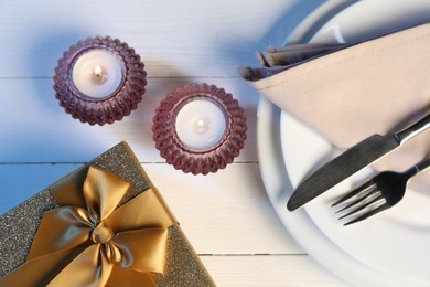 Beautiful table setting with romantic gift and burning candles on white wooden background, flat lay