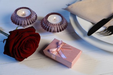 Beautiful table setting with romantic gift, flower and burning candles on white wooden background, closeup