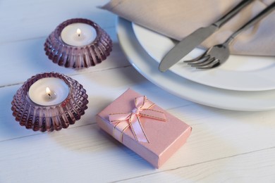 Photo of Beautiful table setting with romantic gift and burning candles on white wooden background, closeup