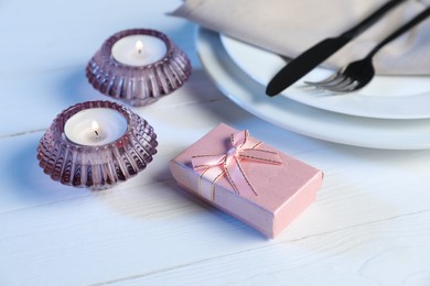 Beautiful table setting with romantic gift and burning candles on white wooden background, closeup