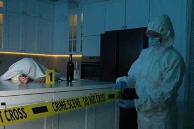 Photo of Forensic expert blocking way to crime scene and dead body with yellow tape indoors