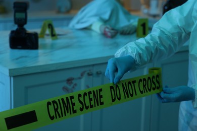 Photo of Forensic expert blocking way to crime scene with yellow tape indoors, closeup