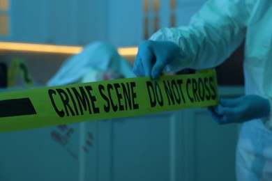 Photo of Forensic expert blocking way to crime scene with yellow tape indoors, closeup