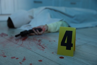 Photo of Dead body with gun at crime scene indoors, focus on numbered marker stand